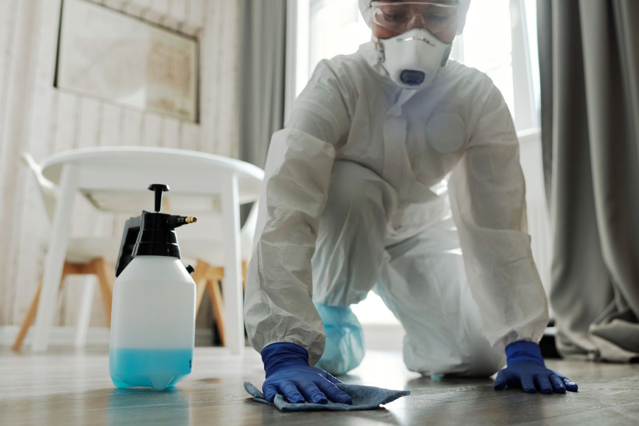 about-img-01 Person in protective gear cleaning a floor with disinfectant spray and cloth indoors.