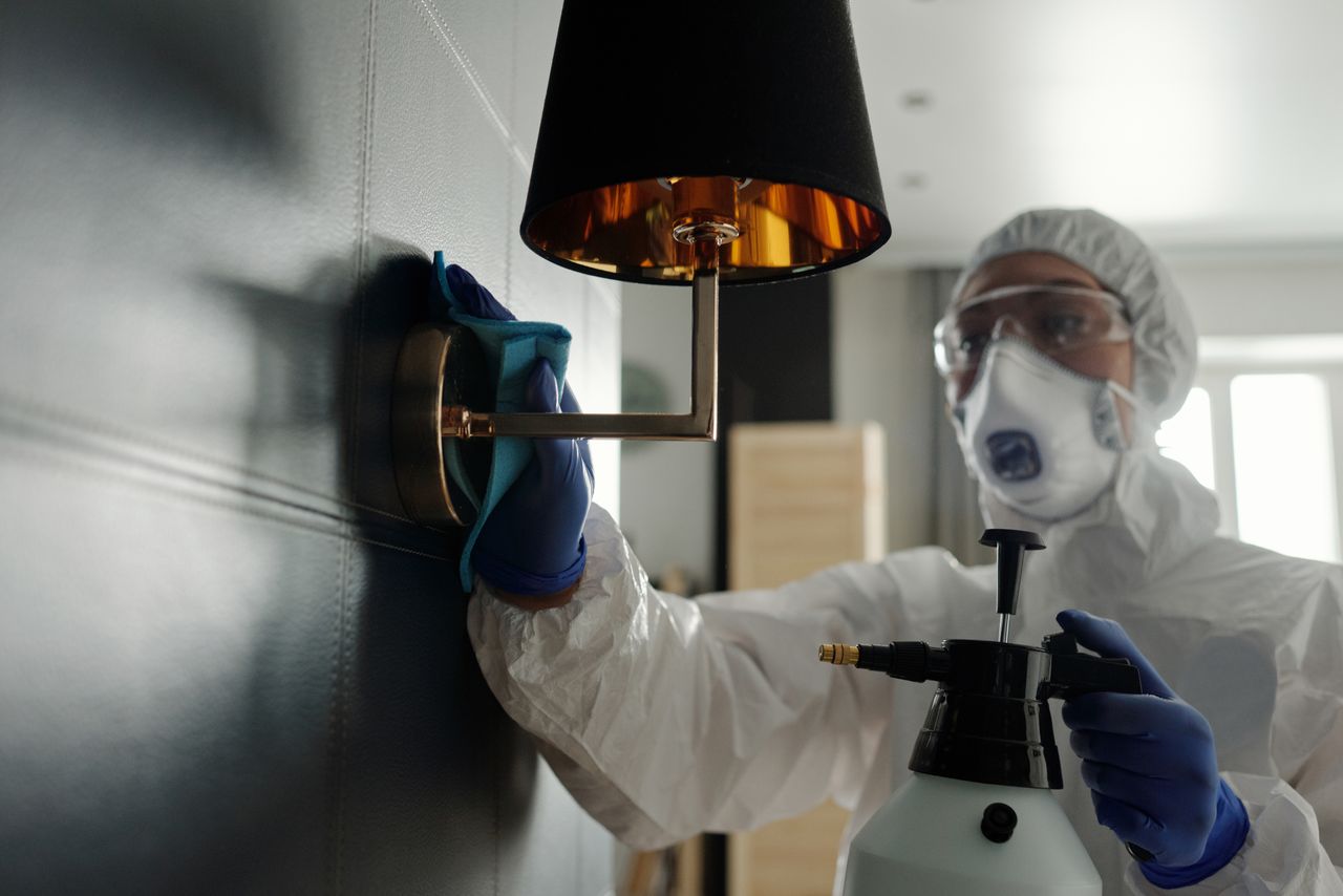 gallery-03 A person wearing protective gear sanitizes a wall lamp to prevent virus spread.